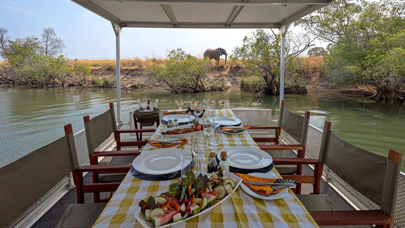 Guests dine on a river deck as an elephant feeds nearby at Ila Safari Lodge.