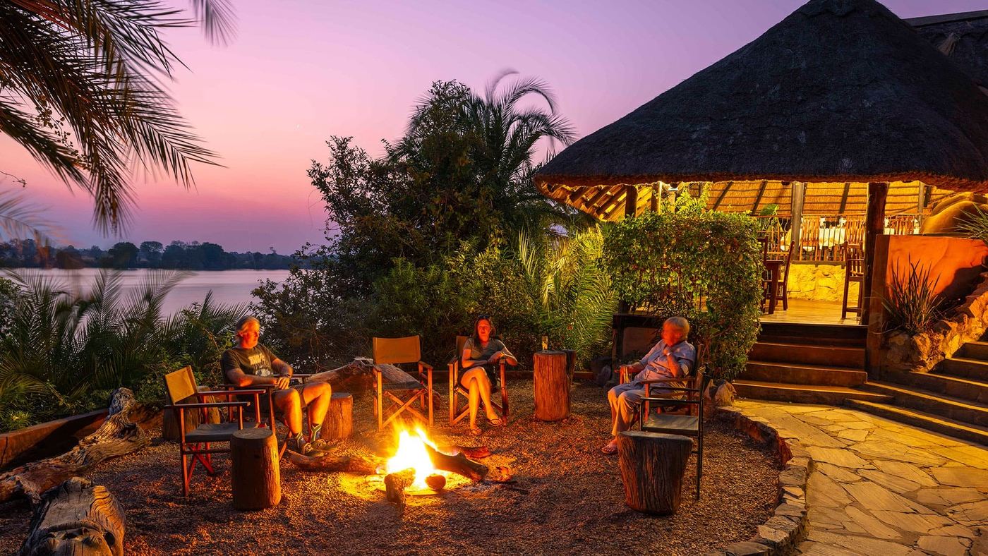 Guests relax by a fire pit at dusk beside the Kafue River at Mukambi Safari Lodge.